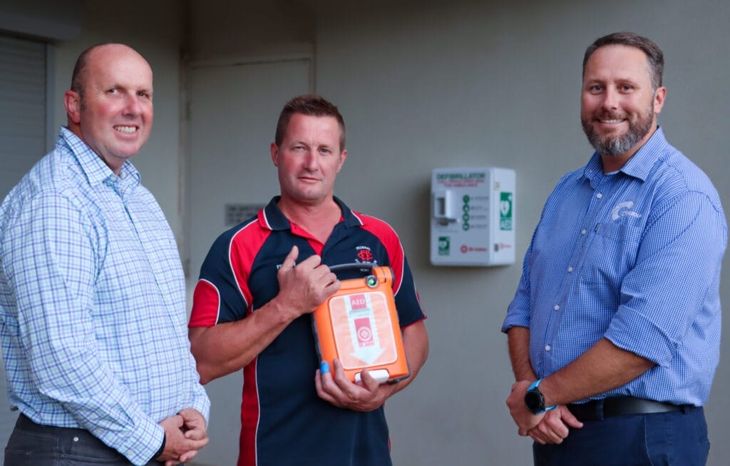 Two men stand next to a another man who is holding an Automated External Defibillator (AED).