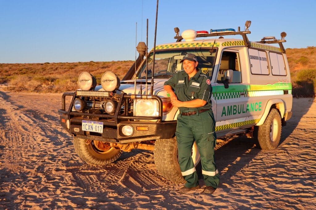 A person in a uniform stands next to an ambulance troop carrier on a beach at sunset.