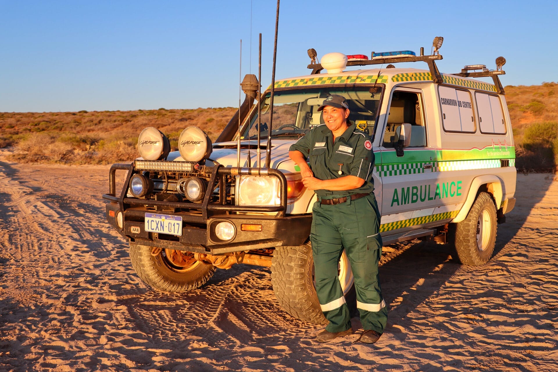A person in a uniform stands next to an ambulance troop carrier on a beach at sunset.
