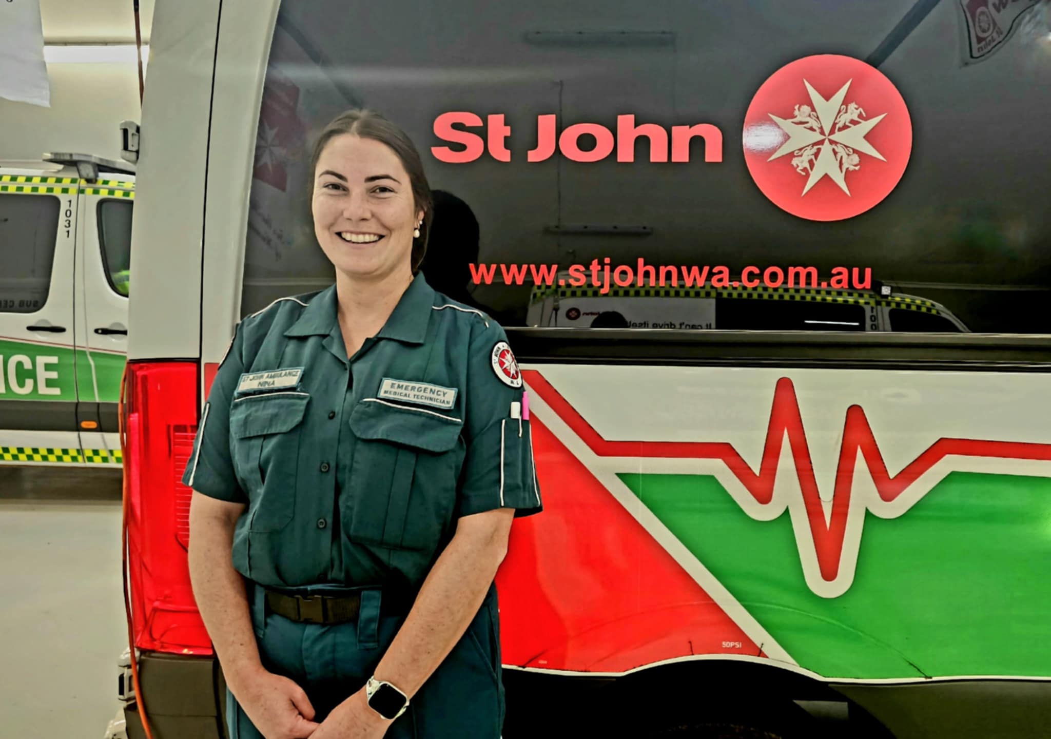 A woman in uniform stands smiling in front of a red, white and green St John WA ambulance.