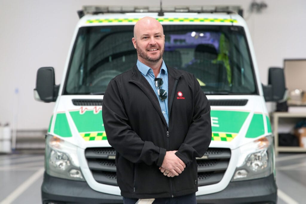 A man in a black jacket stands smiling in front of an ambulance