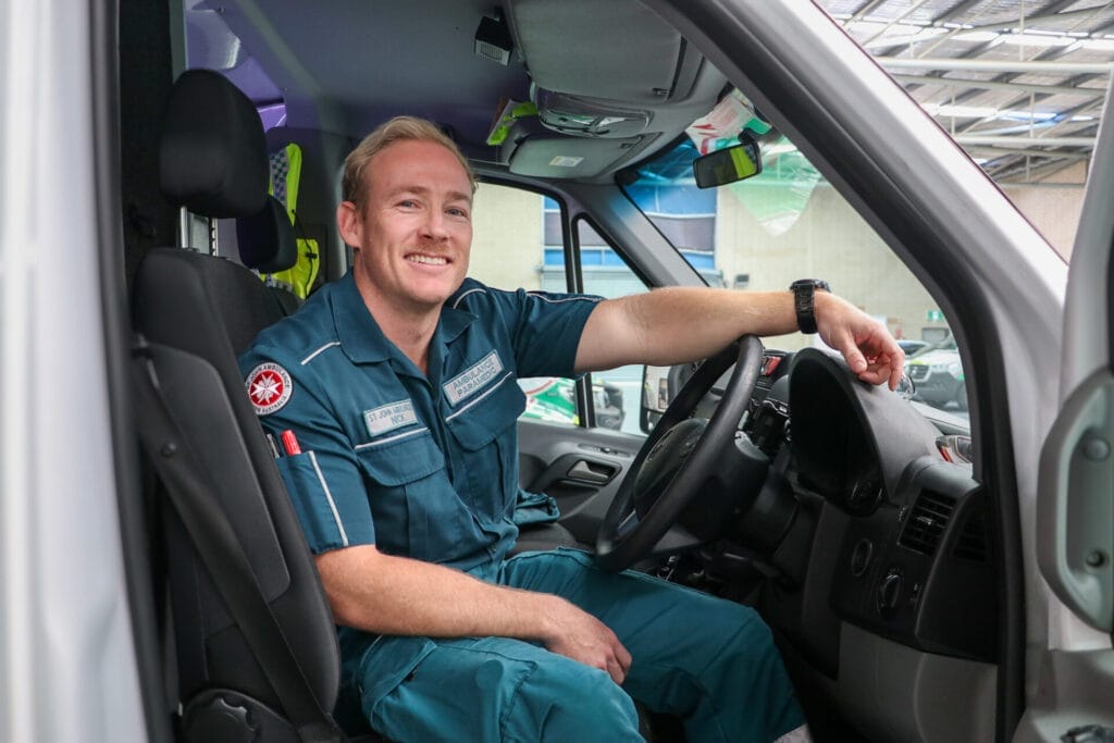 A man in a green uniform sits smiling in the front of an ambulance