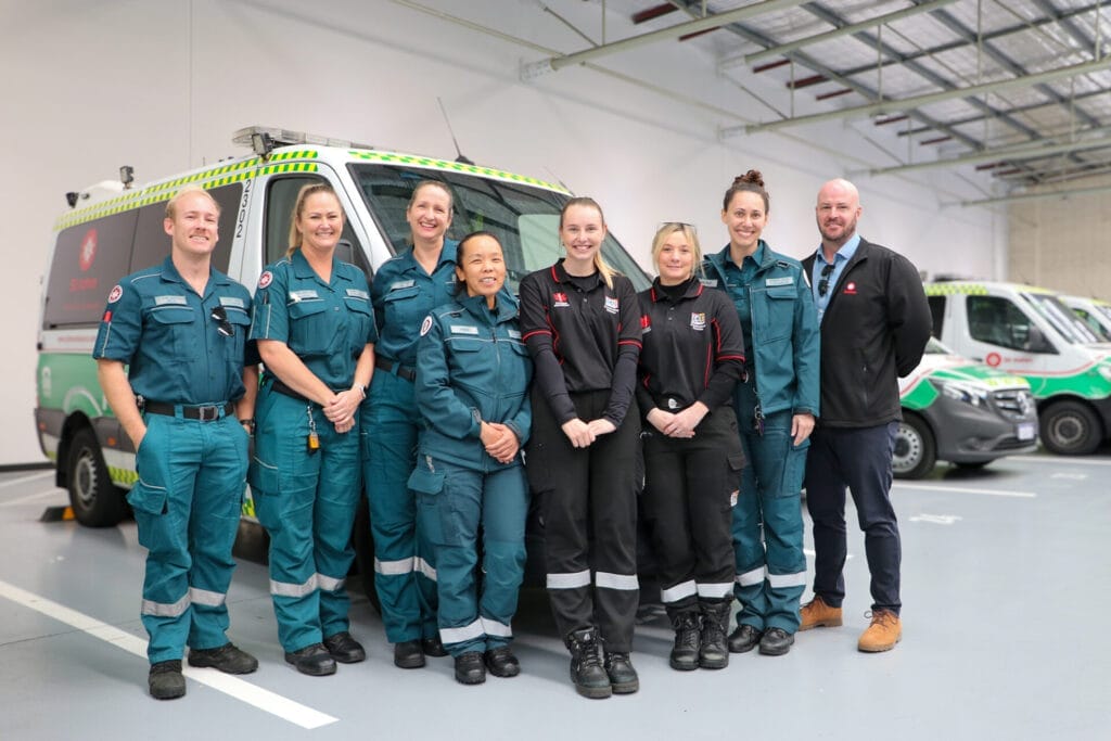 A group of people in green uniforms stand in front of several ambulances inside a depot.