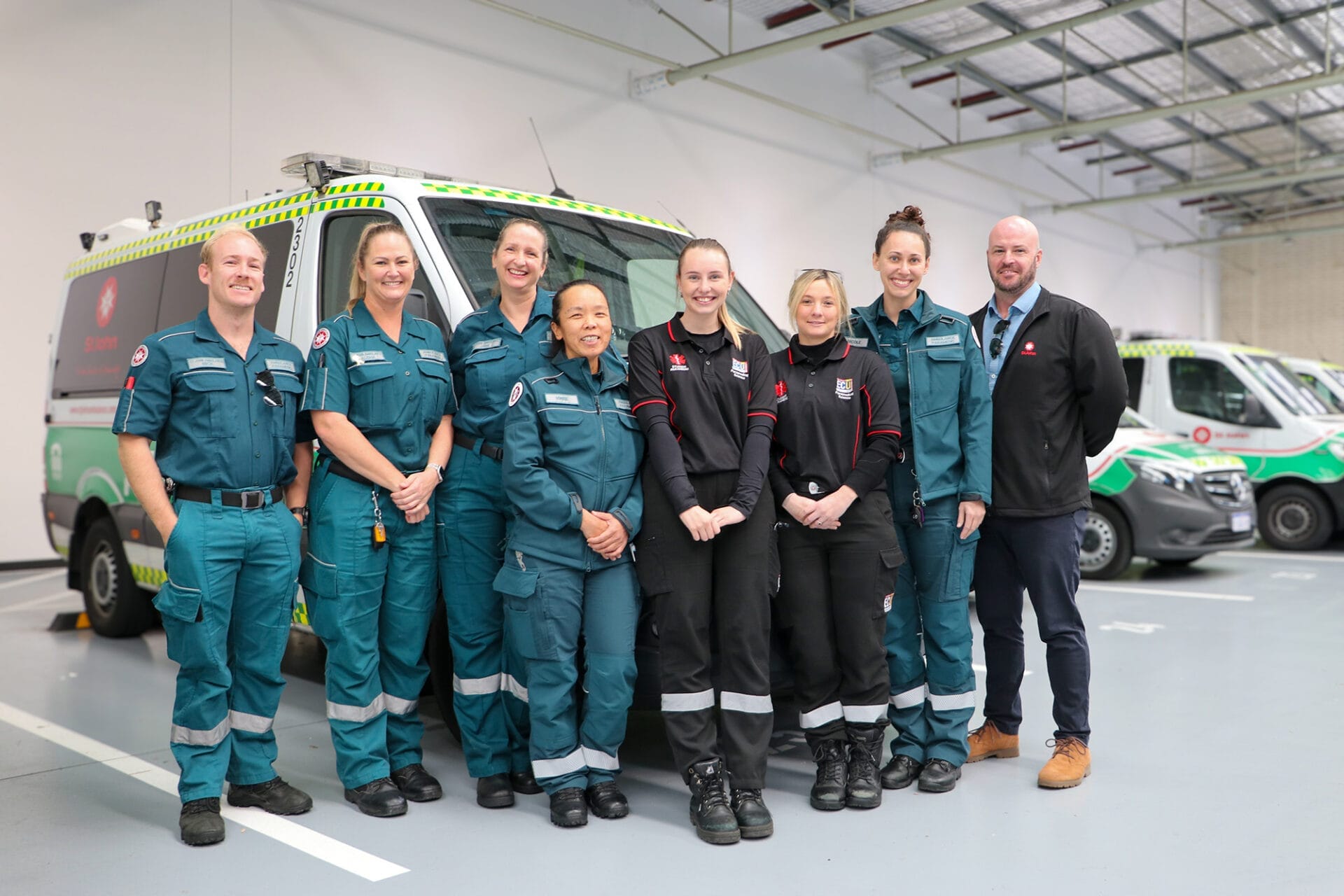 A group of people in green uniforms stand in front of several ambulances inside a depot.