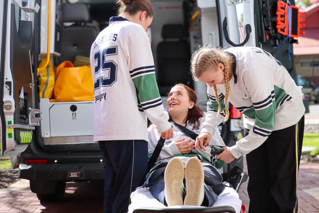 Three students use an automatic stretcher at the back of an ambulance